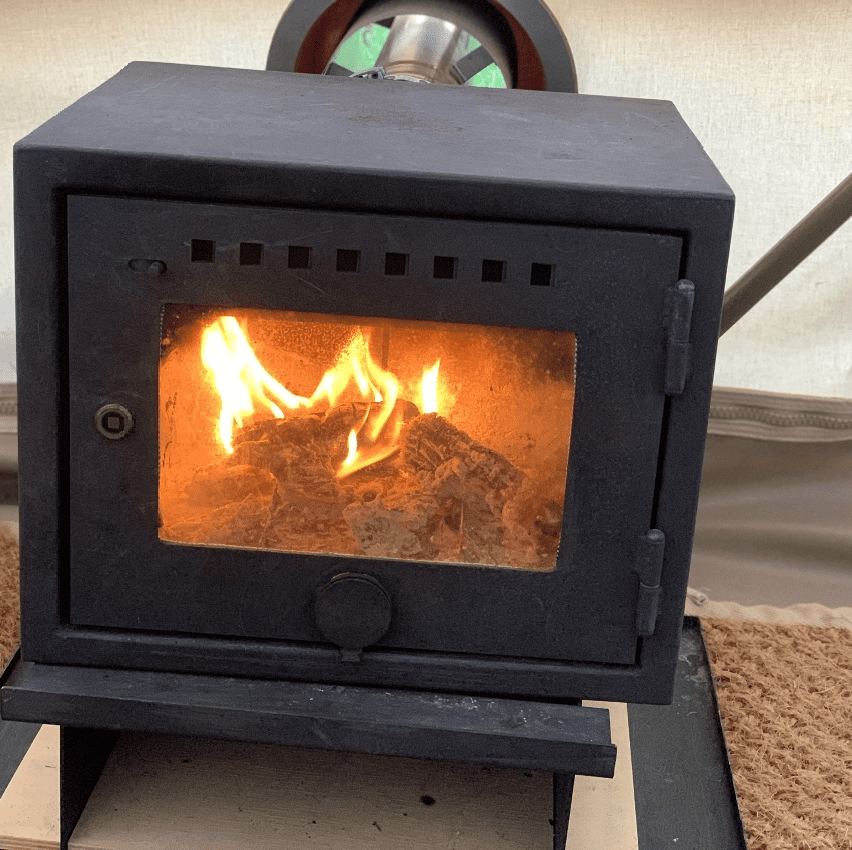 A black wood-burning stove with a glass door and burning logs inside sits on a metal stand.