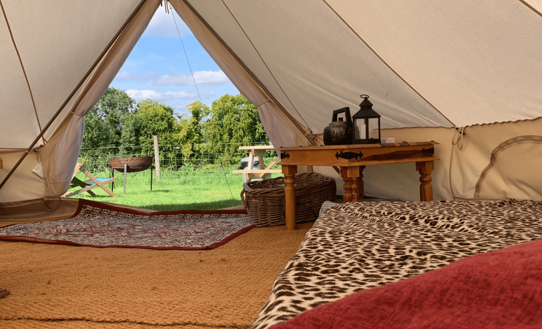 Interior of a canvas tent with a leopard-print bedspread, a wooden table with lanterns, and a view of a grassy campsite with trees outside.