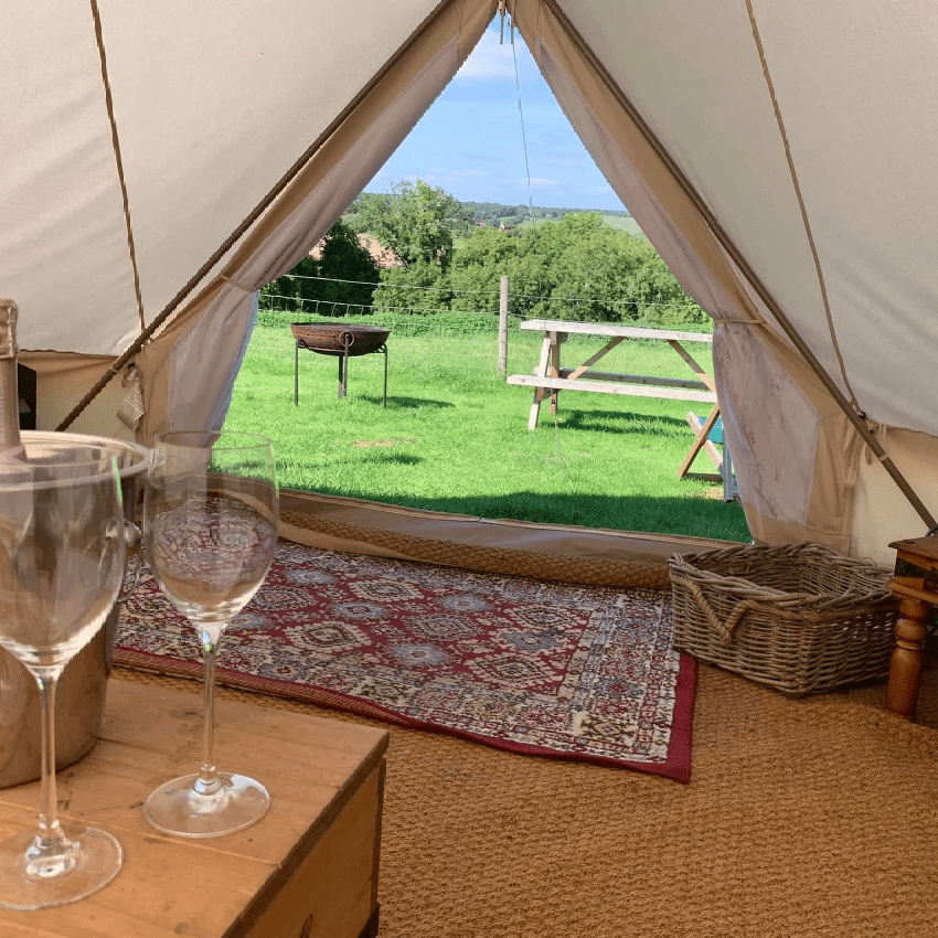 Inside a canvas tent, wine glasses sit on a wooden table beside a patterned rug, with an open flap revealing a grassy yard, a picnic table, and a blue sky.