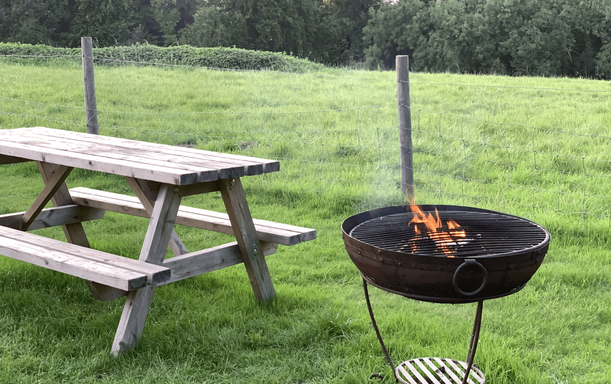 A wooden picnic table sits on a grassy field next to a round charcoal grill with a small fire.