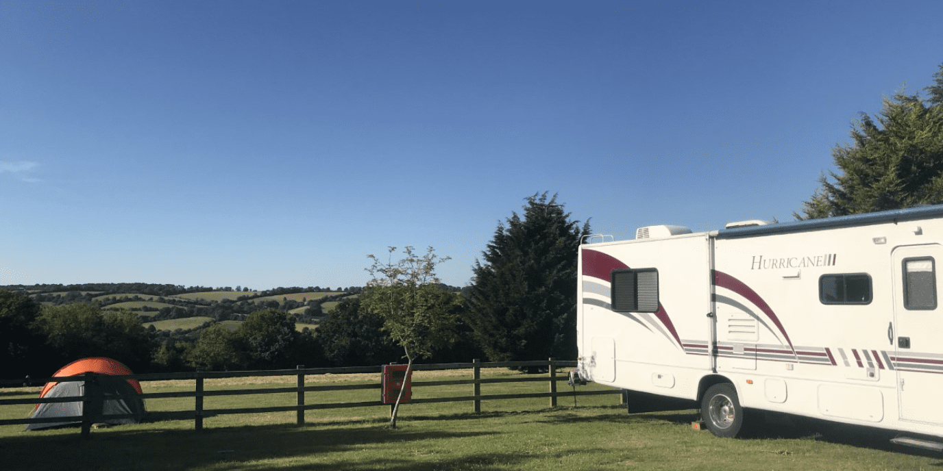A grassy campsite with a large white motorhome on the right, a small tent on the left, and a person standing near a wooden fence with trees and hills in the distance.