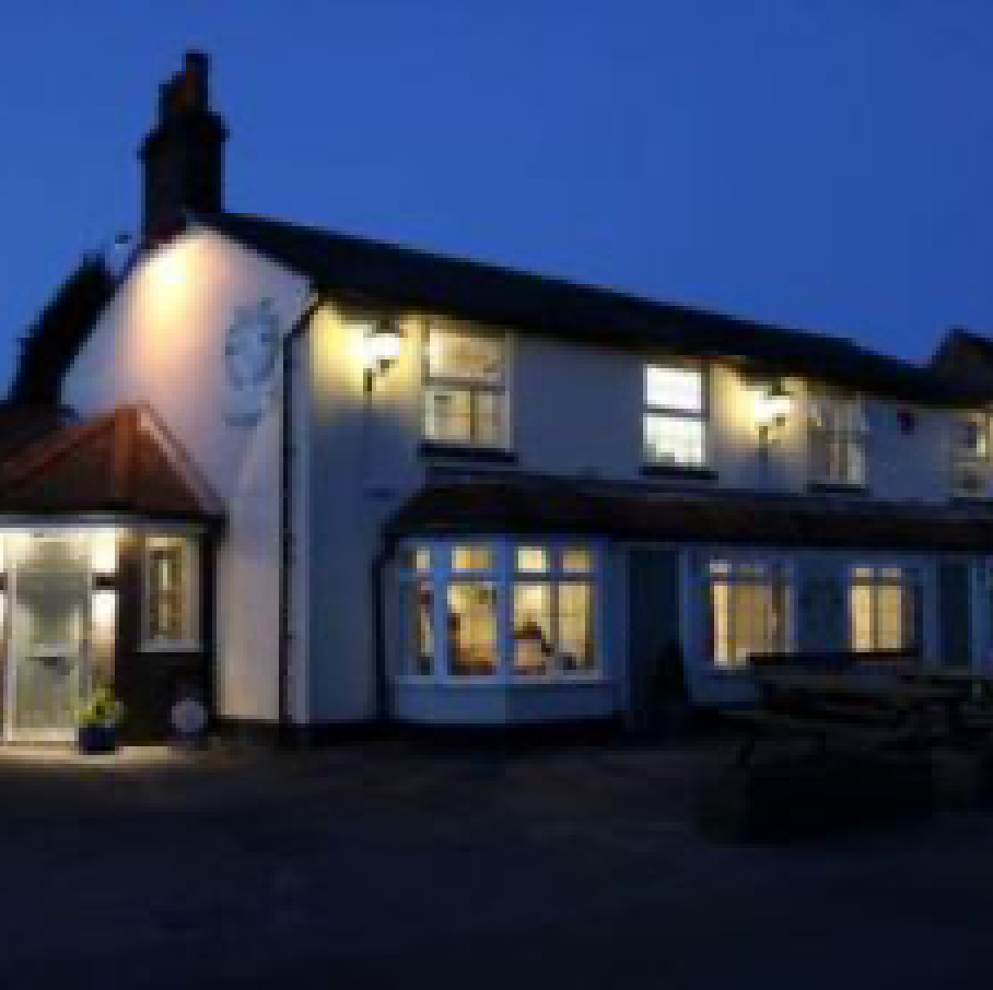 A white two-story pub at night with warm lights in the windows and an outdoor bench by the entrance.