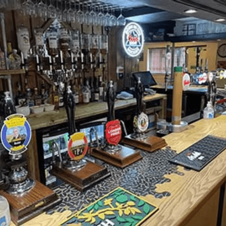 Bar counter lined with numerous beer taps and beer mats, with bottles displayed on the shelves behind.