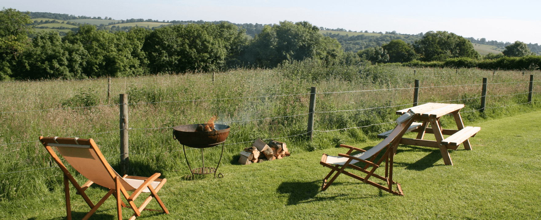 A sunny grassy backyard with wooden lounge chairs, a small fire pit, and a picnic table beside a wooden fence with distant trees.