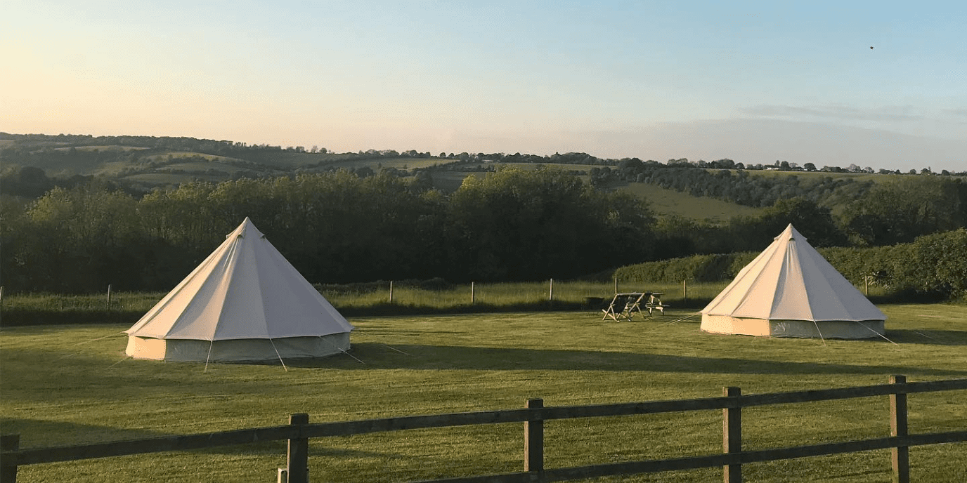 Two white canvas tents pitched on a grassy field with a wooden fence in the foreground and a tree-lined horizon under a clear sky.
