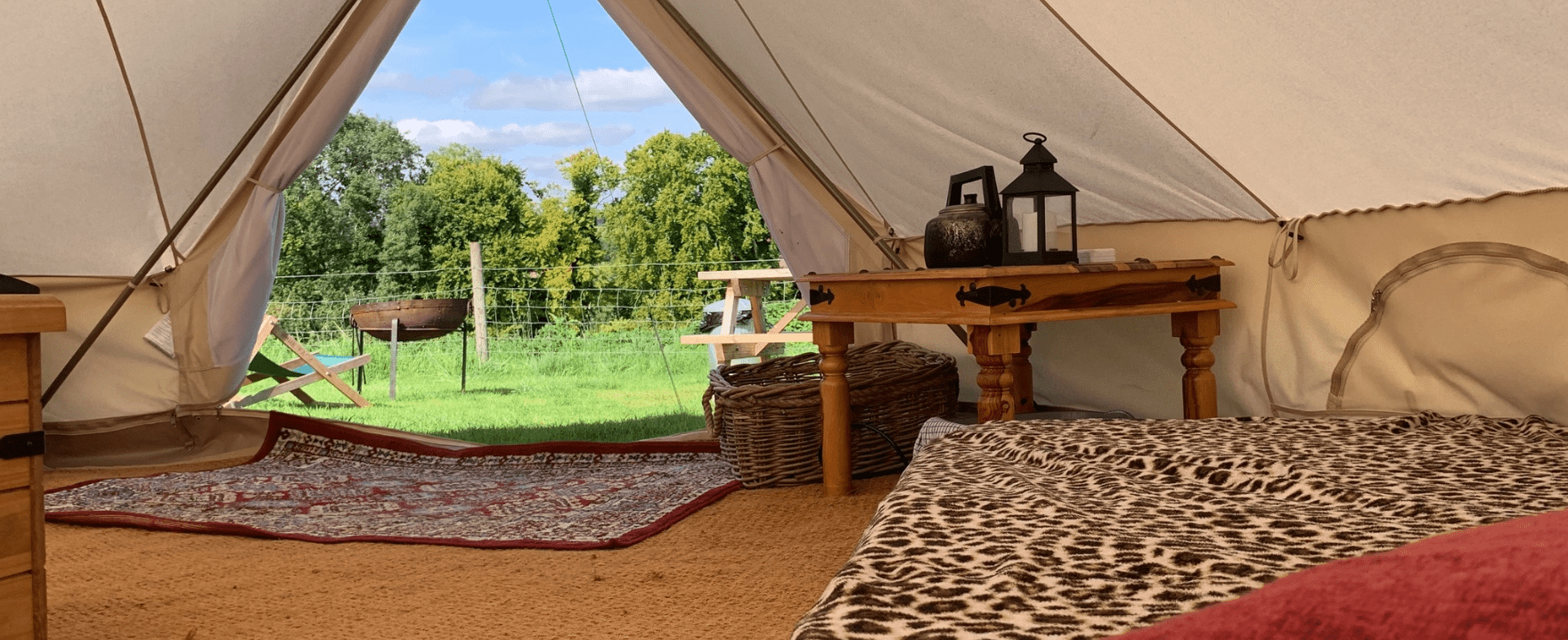 interior of glamping tent with wooden table and rug and a view outside the entrance showing benches and a fire pit