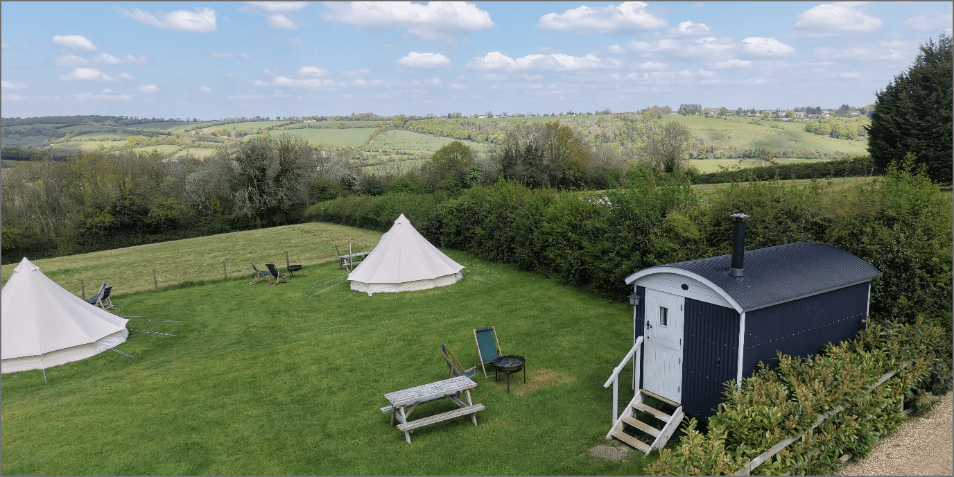 A grassy campsite featuring two white tents, a dark caravan, and outdoor seating on a neatly mowed field with hedges and distant rolling hills.