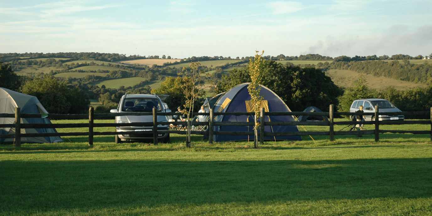A rural campsite with tents and parked cars behind a wooden fence on a grassy field, with rolling hills and a blue sky in the background.