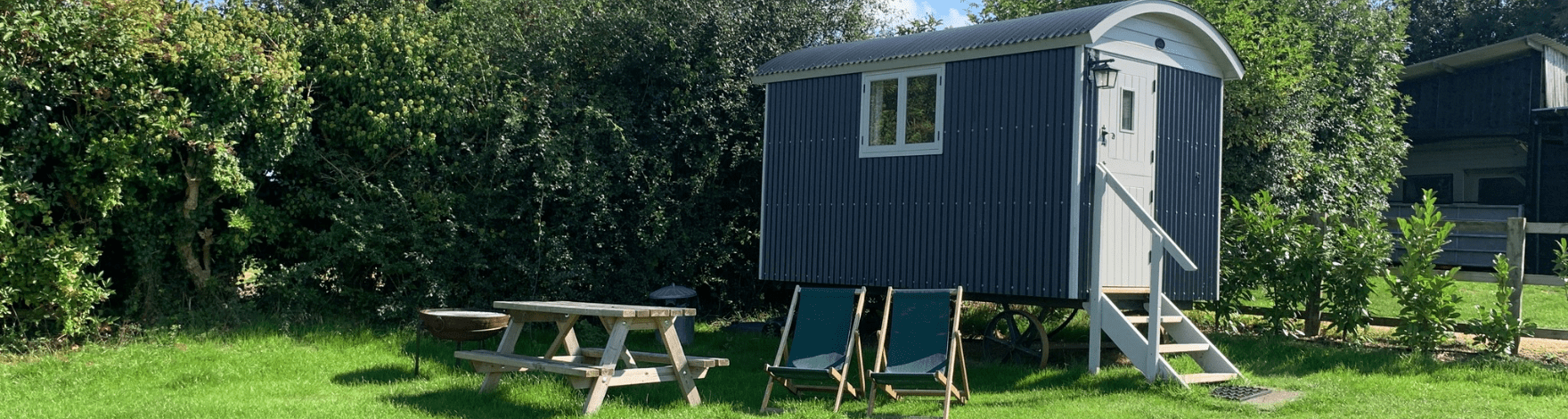 exterior view of a camping hut with wooden bench, fire pit, and two deck chairs positioned in front of a tall hedgerow