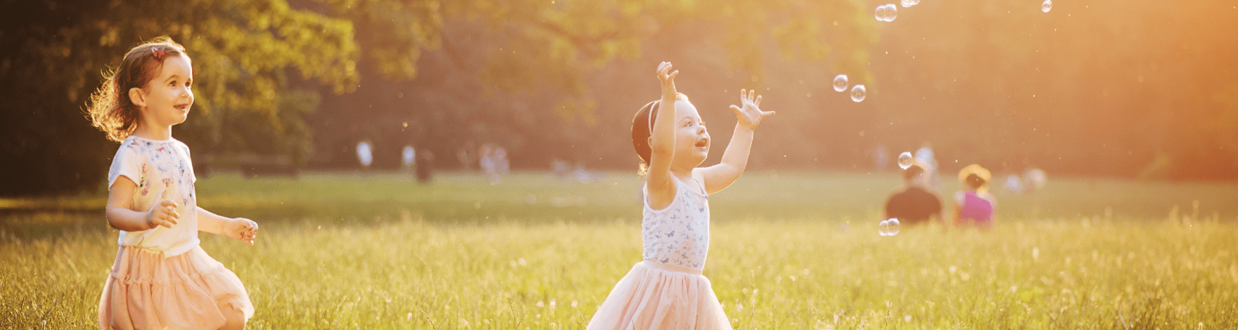 two children playing in a field chasing bubbles, people sitting down in the field in the background out of focus