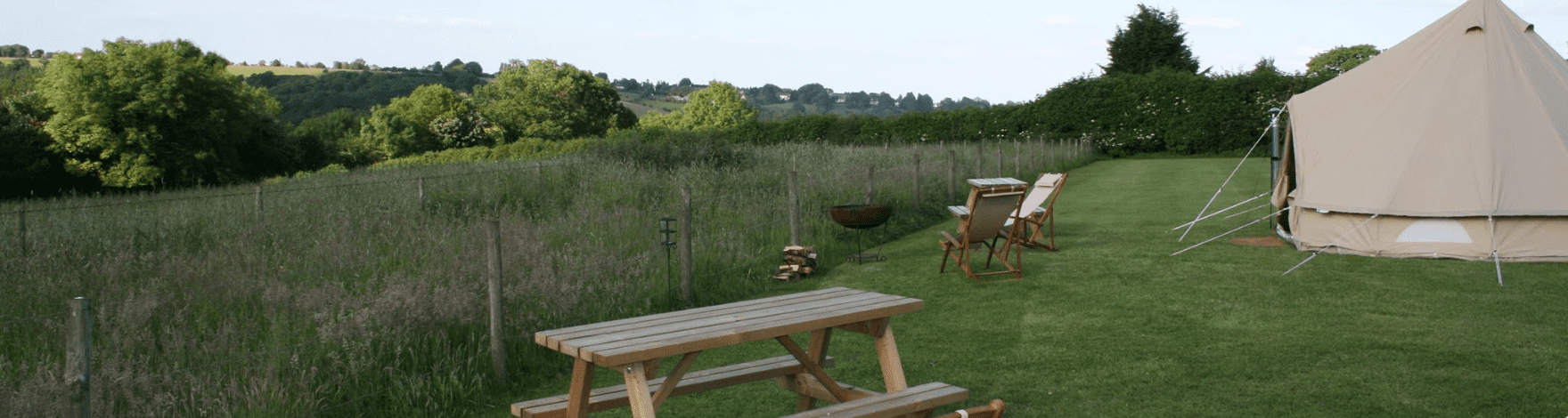 rural view of a glamping tent, two wooden benches, two deck chairs, fire pit, log pile, and fence with the rolling countryside hills behind