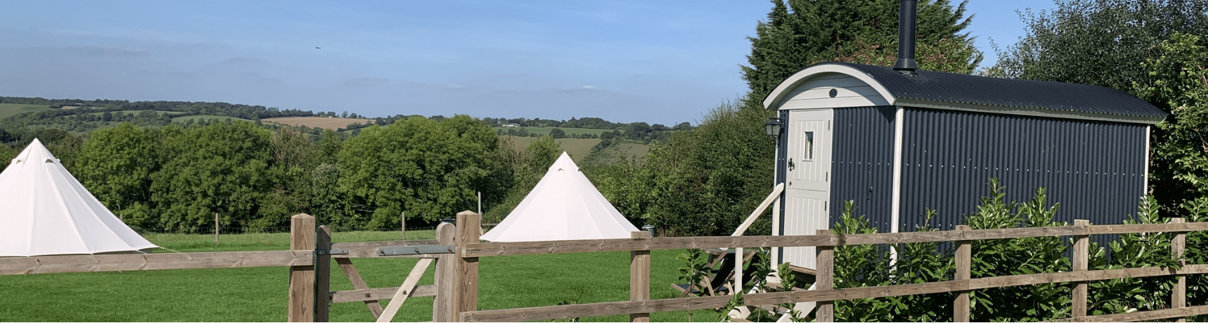 two glamping tents and a camping hut building in the rural countryside behind a wooden fence and small wooden gate