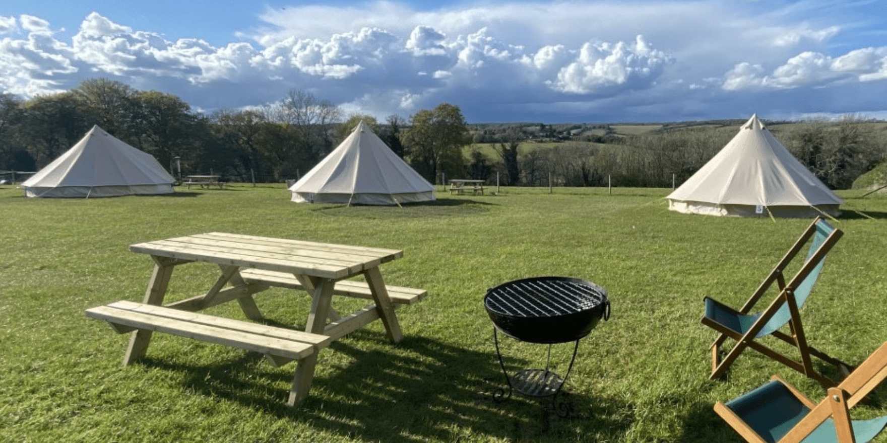 A grassy campsite with three beige canvas tents, a wooden picnic table, a small round metal fire pit, and two wooden lounge chairs under a blue, partly cloudy sky.