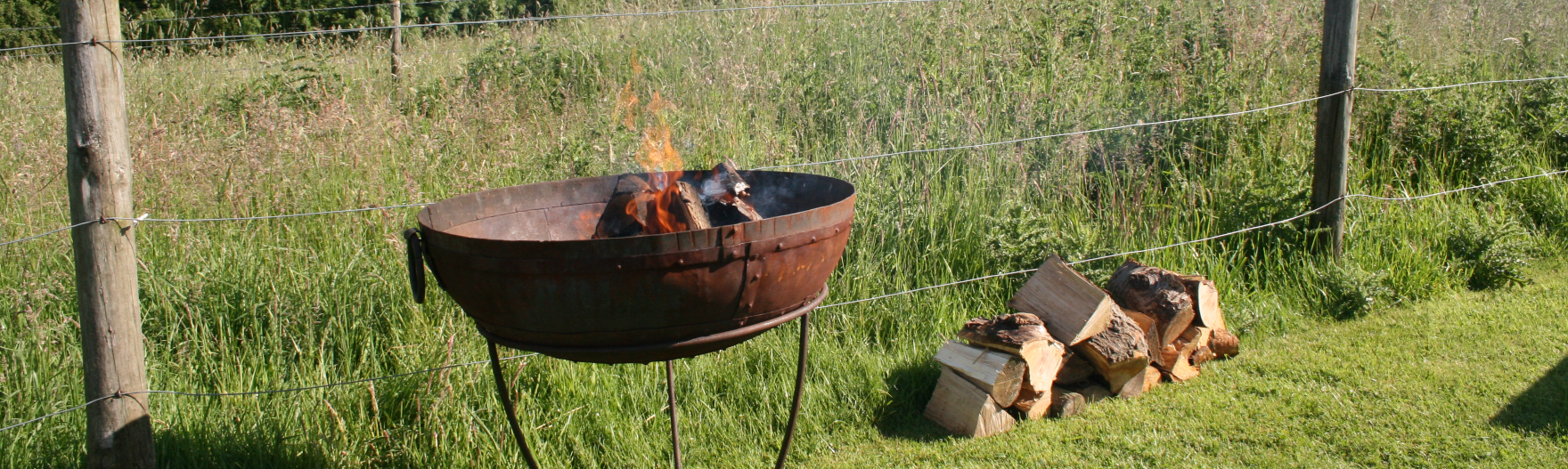 a fire pit with a fire going and a pile of logs beside located in the edge of a field by a fence with long grass behind