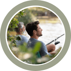 Two men fishing by a lake with greenery around.