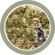 A child wearing a helmet and harness walks along a wooden rope bridge in a forest.